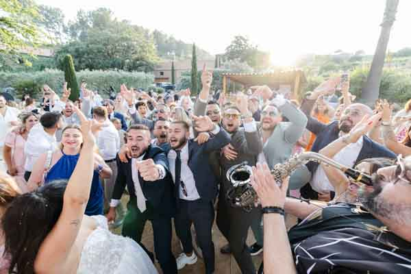 Groupe d'invités et mariés célébrant avec un saxophoniste lors d'un cocktail en extérieur.