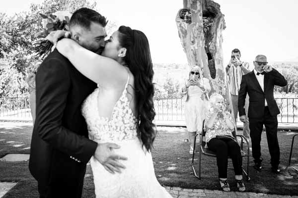 Photographie en noir et blanc d'un baiser passionné entre les mariés lors d'une cérémonie en extérieur.