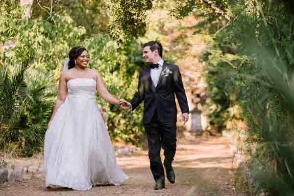 Couple de mariés marchant main dans la main dans un domaine arboré en Provence.