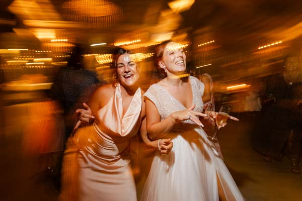 Photo festive avec effet de mouvement montrant la mariée et une amie dansant avec un verre à la main.