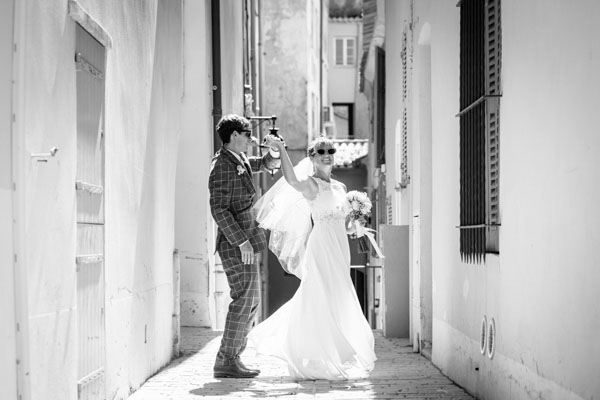 Photographie en noir et blanc d'un couple de mariés dansant dans une ruelle typique d'un village provençal.