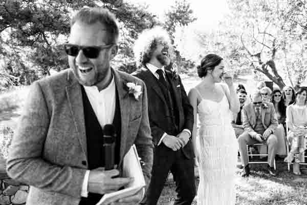 Un homme avec un micro rit aux éclats pendant un discours de mariage en extérieur, entouré d'invités émus.