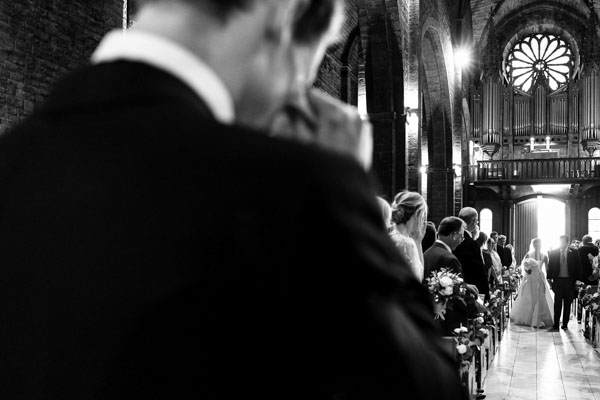 Vue en noir et blanc depuis l'autel de la mariée entrant dans une église historique au bras de son père.