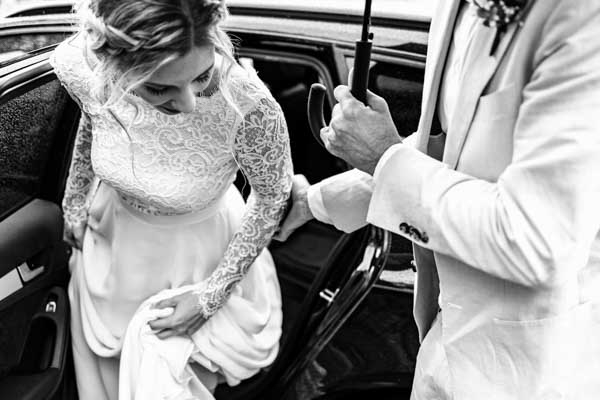 Gros plan en noir et blanc d'une mariée en robe de dentelle montant en voiture sous un parapluie.