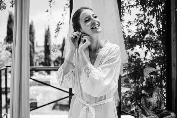 Portrait en noir et blanc d'une mariée souriante ajustant ses boucles d'oreilles avant d'enfiler sa robe.