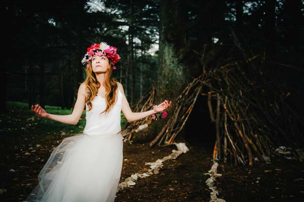 Portrait d'une mariée bohème avec couronne de fleurs, debout dans une forêt de Provence avec un tipi en branches.