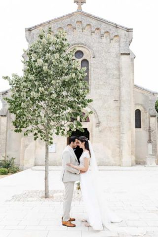 Couple de mariés s'embrassant tendrement devant la façade d'une église en pierre de Provence.
