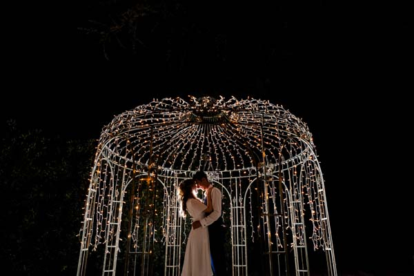 Silhouette d'un couple de mariés s'embrassant sous une arche de guirlandes lumineuses la nuit.