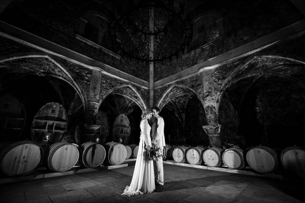 Couple de mariés posant dans une cave de domaine viticole entouré de fûts de chêne, en noir et blanc.