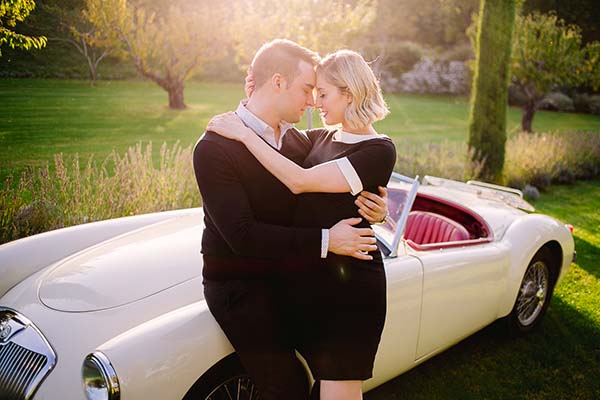 Couple de futurs mariés s'enlaçant tendrement devant une voiture ancienne blanche dans le jardin de l'Abbaye Sainte Eusèbe dans le Vaucluse.