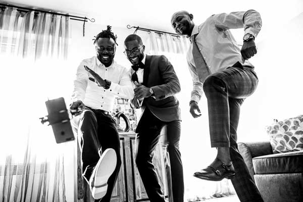 Trois hommes, dont le marié, se penchant de manière ludique devant une caméra de téléphone sur un trépied, s'amusant pendant les préparatifs.