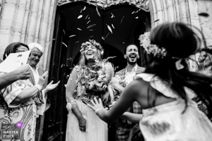 Photographie de mariage primée en noir et blanc montrant des mariés riant aux éclats sous un lancer de feuilles d'olivier à la sortie d'une église en Provence.