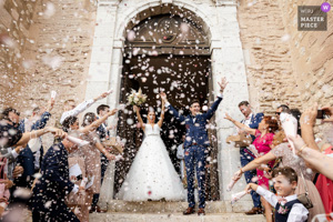 Photographie de mariage primée montrant des mariés radieux levant les bras sous une pluie de pétales de fleurs lors de leur sortie d'église en Provence.