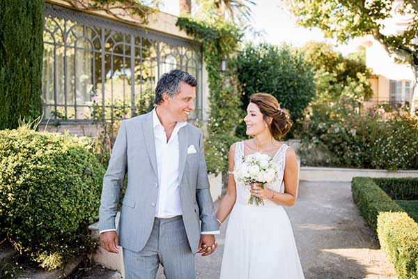 Couple de mariés complices marchant main dans la main dans l'allée d'un jardin provençal à Saint-Tropez.