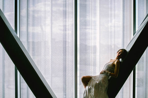 Mariée posant contre une structure métallique devant une grande baie vitrée lumineuse.