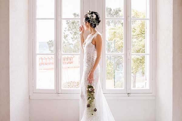 Portrait de profil d'une mariée en robe de dentelle blanche tenant un bouquet cascade devant une fenêtre lumineuse.