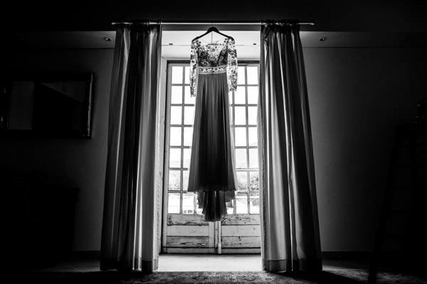 Robe de mariée en dentelle suspendue devant une fenêtre lumineuse en noir et blanc.