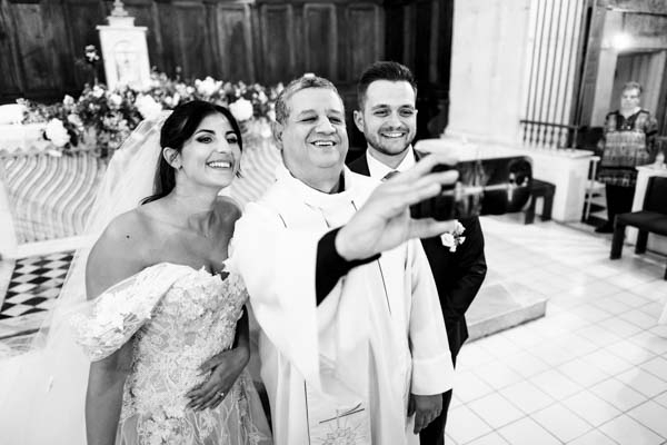 Photo noir et blanc d'un prêtre prenant un selfie avec les mariés à l'autel.