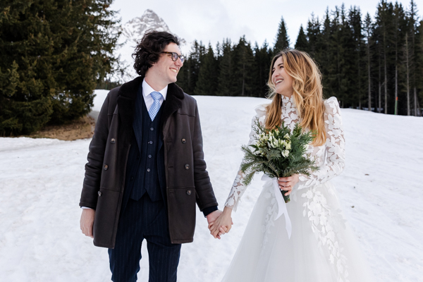 Couple de mariés complices marchant dans la neige devant une forêt de sapins à Méribel.