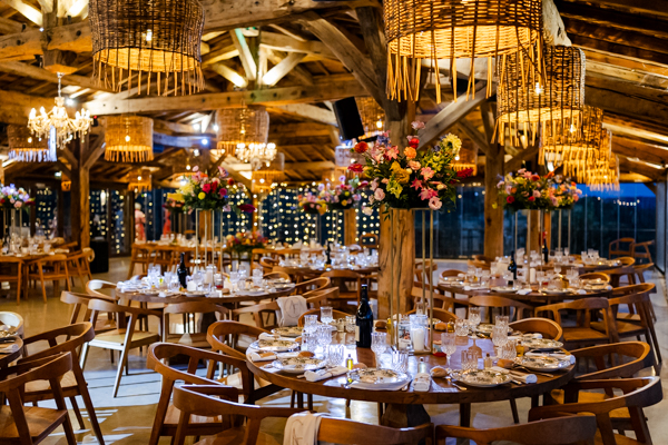 Salle de réception du Domaine de Brès avec tables rondes en bois, charpente apparente et suspensions en rotin pour un mariage bohème chic.