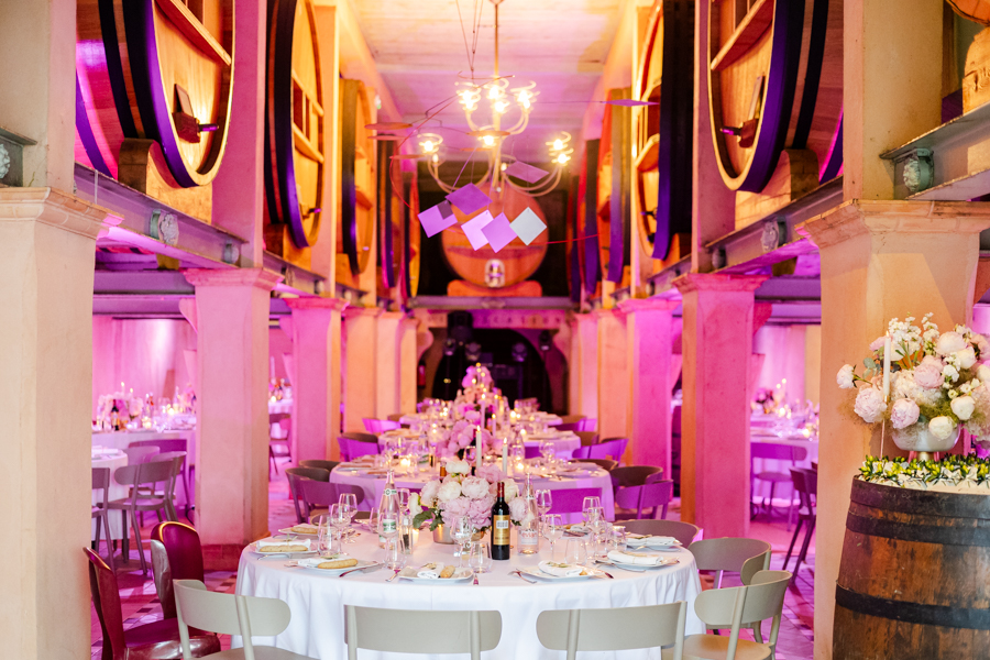 Salle de réception de mariage installée dans le chai du Domaine Bunan, éclairée en rose, avec les grands foudres en arrière-plan.