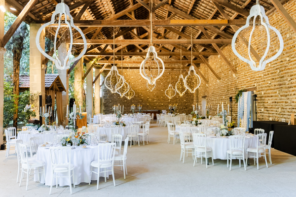Salle de réception du mariage dans la grange en pierre du Château de Vergières, décorée avec des tables rondes et de grands lustres blancs.