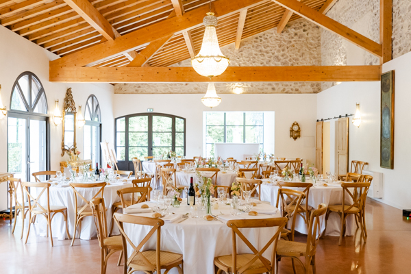 Salle de réception du Domaine de Romanil dressée pour un dîner de mariage, avec chaises en bois, lustres et murs en pierre apparente.