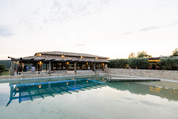 Ambiance de soirée au bord de la piscine du Domaine de Brès, avec le pool house illuminé se reflétant dans l'eau au crépuscule.