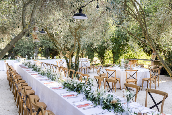 Longue table de réception de mariage dressée en extérieur sous les oliviers à la Bastide du Régent, décorée de fleurs blanches.